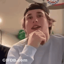 A Young Man Wearing A Baseball Cap And A White Sweater Is Sitting In A Living Room With His Hands On His Chin . GIF