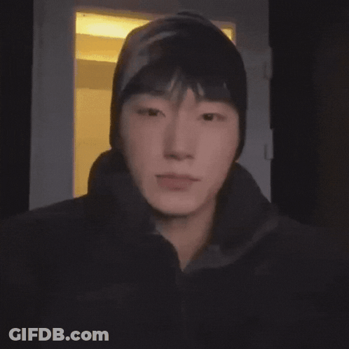 A Young Man Wearing A Beanie Looks At The Camera In A Dark Room GIF
