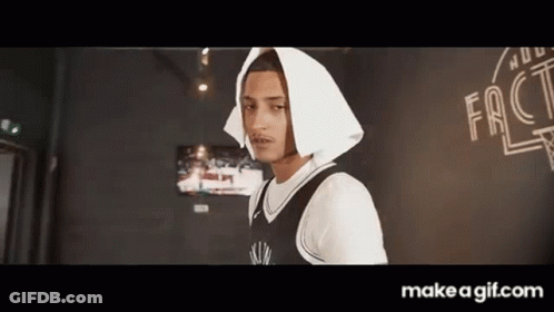 A Young Man Wearing A Black And White Basketball Jersey And A White Hooded Jacket Is Standing In A Room . GIF