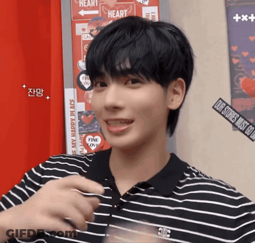 A Young Man Wearing A Black And White Striped Shirt Is Standing In Front Of A Wall With Stickers On It That Say Heart GIF