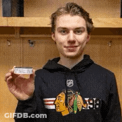 A Young Man Wearing A Black Chicago Blackhawks Hoodie Is Holding A Small Piece Of Paper . GIF
