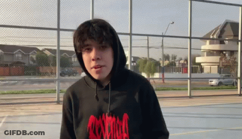 A Young Man Wearing A Black Hoodie And A Red Shirt Is Standing In Front Of A Fence . GIF