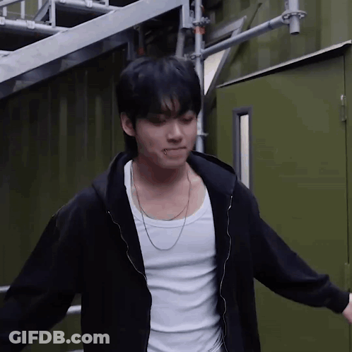 A Young Man Wearing A Black Hoodie And A White Tank Top Is Standing In Front Of A Green Building . GIF