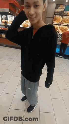 A Young Man Wearing A Black Hoodie And Grey Sweatpants Is Standing In Front Of A Fast Food Restaurant GIF