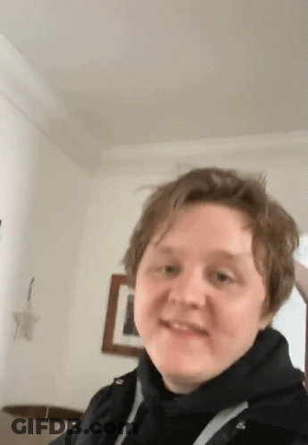 A Young Man Wearing A Black Hoodie Is Standing In A Living Room . GIF