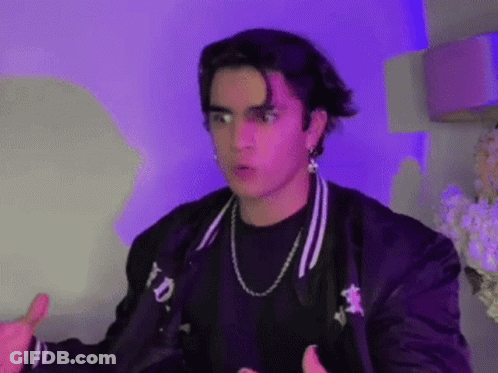 A Young Man Wearing A Black Jacket And A Necklace Is Giving A Thumbs Up Sign . GIF