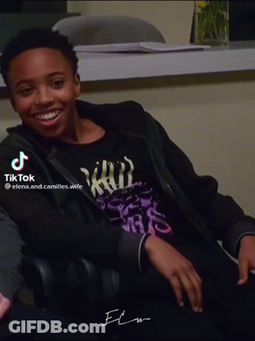 A Young Man Wearing A Black Jacket And A Purple Shirt Is Smiling And Sitting In A Chair GIF