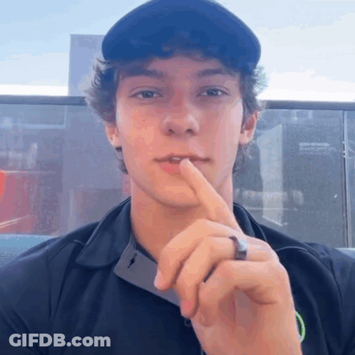 A Young Man Wearing A Black Shirt And A Black Hat Holds His Finger To His Lips GIF