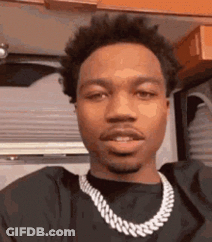 A Young Man Wearing A Black Shirt And A Chain Around His Neck Is Looking At The Camera . GIF