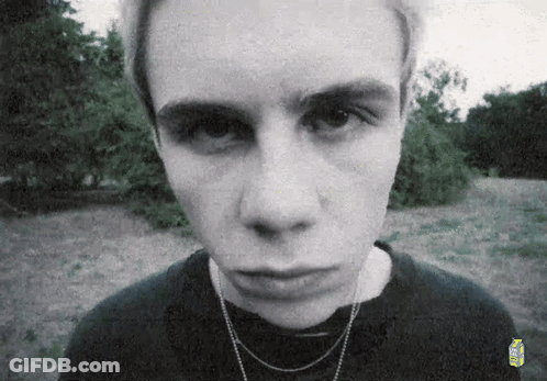A Young Man Wearing A Black Shirt And A Necklace Looks At The Camera . GIF