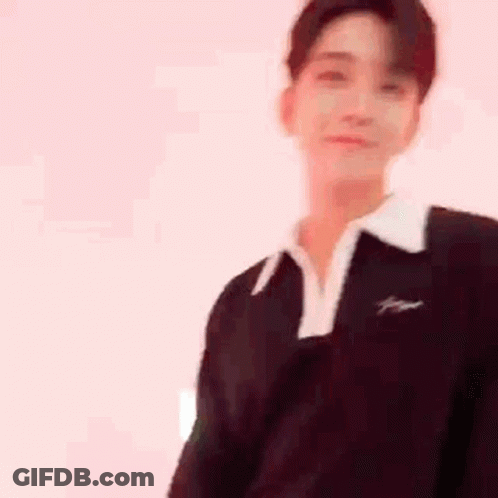 A Young Man Wearing A Black Sweater And A White Collar Is Standing In Front Of A White Wall . GIF