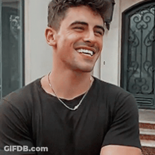A Young Man Wearing A Black T-Shirt And A Necklace Is Smiling . GIF