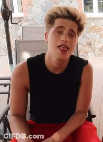 A Young Man Wearing A Black Tank Top And Red Shorts Is Sitting In A Chair GIF