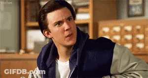 A Young Man Wearing A Blue And Gray Jacket Is Sitting In A Library . GIF