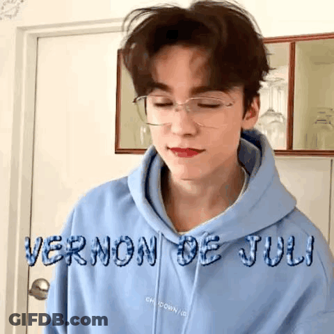 A Young Man Wearing A Blue Hoodie And Glasses Is Standing In Front Of A Door . GIF