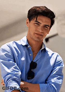 A Young Man Wearing A Blue Shirt And Sunglasses Is Standing With His Arms Crossed And Smiling . GIF
