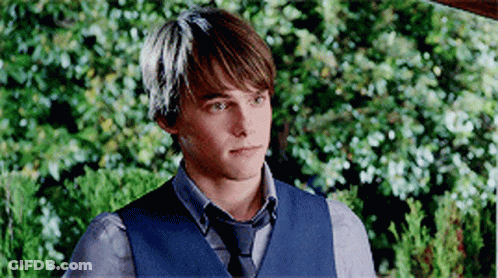 A Young Man Wearing A Blue Vest And Tie Is Standing In Front Of A Bush . GIF