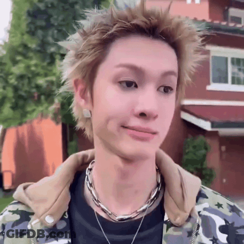 A Young Man Wearing A Camouflage Jacket And A Necklace Looks At The Camera GIF