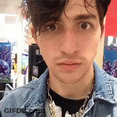 A Young Man Wearing A Denim Jacket And A Necklace Looks At The Camera GIF