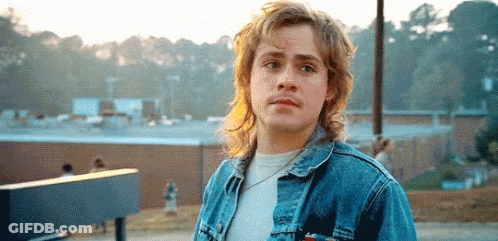 A Young Man Wearing A Denim Jacket And A White Shirt Is Standing In Front Of A Building . GIF