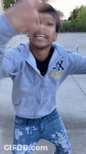 A Young Man Wearing A Gray Hoodie And Blue Jeans Is Standing On A Sidewalk . GIF