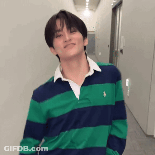 A Young Man Wearing A Green And Blue Striped Shirt Is Standing In A Hallway With His Hands In His Pockets . GIF