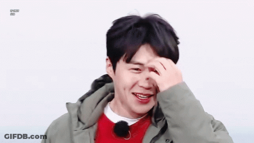 A Young Man Wearing A Green Jacket And A Red Sweater Is Smiling And Covering His Face With His Hand . GIF