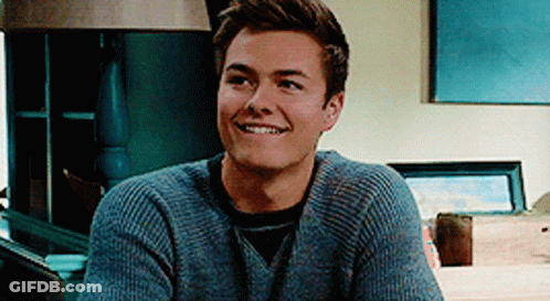 A Young Man Wearing A Grey Sweater Is Smiling While Sitting In Front Of A Lamp . GIF