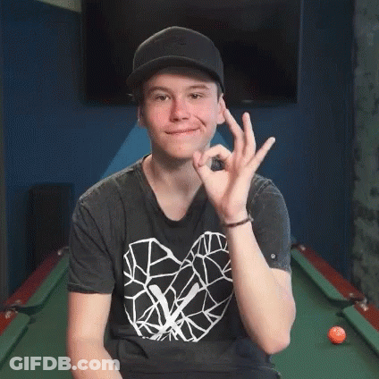 A Young Man Wearing A Hat And A Black Shirt Is Sitting In Front Of A Pool Table Giving An Ok Sign . GIF