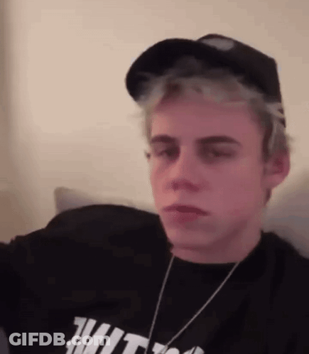 A Young Man Wearing A Hat And A Black Shirt Is Sitting On A Couch . GIF