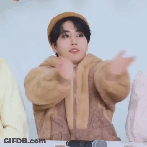 A Young Man Wearing A Hat And A Brown Jacket Is Sitting At A Table . GIF