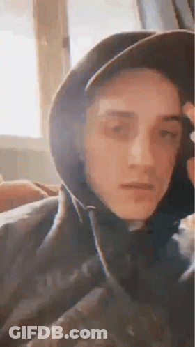 A Young Man Wearing A Hat And A Hoodie Is Sitting In Front Of A Window . GIF