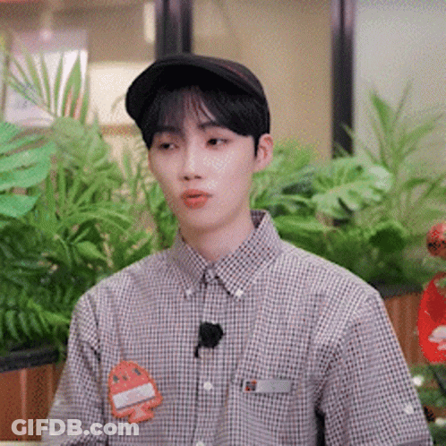 A Young Man Wearing A Hat And A Plaid Shirt Is Sitting In Front Of A Plant . GIF