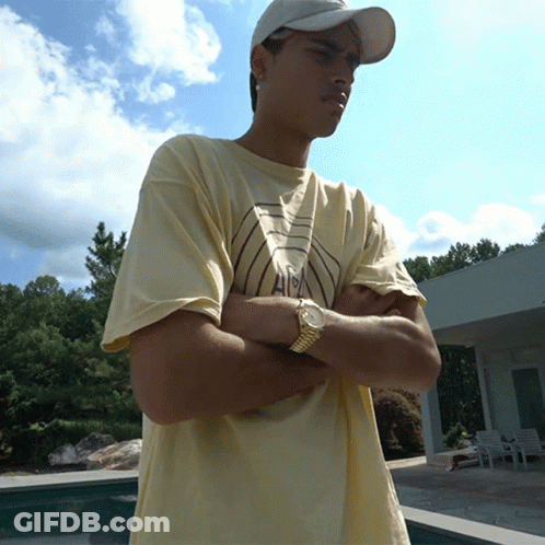 A Young Man Wearing A Hat And A Watch Stands With His Arms Crossed In Front Of A Pool GIF