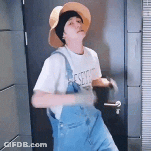 A Young Man Wearing A Hat And Overalls Is Dancing In Front Of A Door . GIF