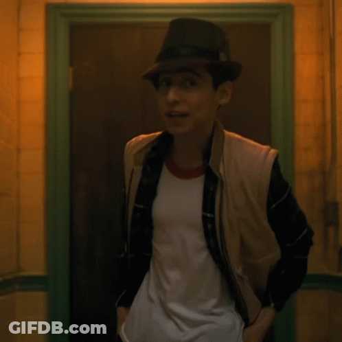 A Young Man Wearing A Hat And Vest Is Standing In A Doorway GIF
