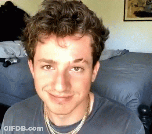 A Young Man Wearing A Necklace And A Gray Shirt Smiles For The Camera GIF