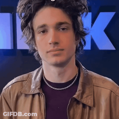 A Young Man Wearing A Necklace And A Leather Jacket Looks At The Camera GIF