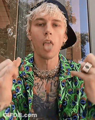 A Young Man Wearing A Peacock Feather Shirt And A Hat Is Sticking His Tongue Out . GIF