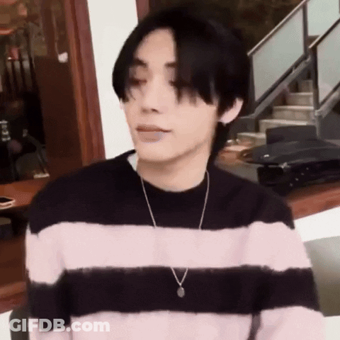 A Young Man Wearing A Pink And Black Striped Sweater And A Necklace Is Sitting In Front Of A Staircase . GIF