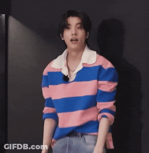 A Young Man Wearing A Pink And Blue Striped Shirt Is Standing In Front Of A Black Wall With His Mouth Open . GIF