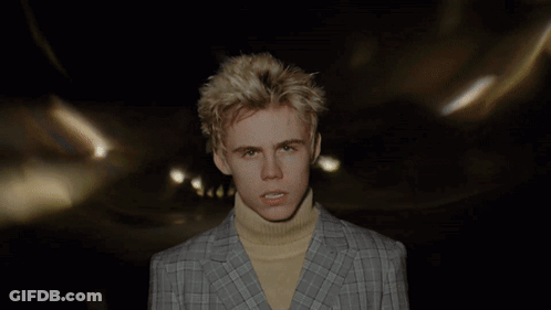 A Young Man Wearing A Plaid Jacket And A Yellow Turtleneck Looks At The Camera GIF