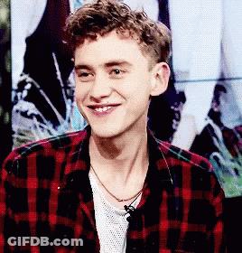 A Young Man Wearing A Plaid Shirt And A Necklace Is Smiling GIF