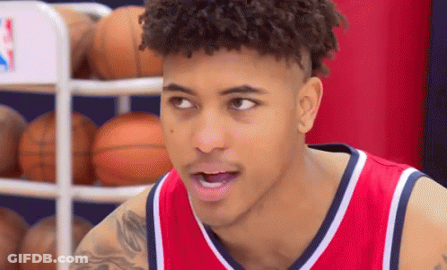 A Young Man Wearing A Red And White Basketball Jersey Is Standing In Front Of A Basketball Rack . GIF