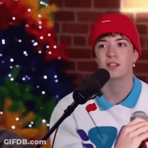 A Young Man Wearing A Red Beanie Is Singing Into A Microphone While Sitting In Front Of A Christmas Tree . GIF