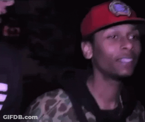 A Young Man Wearing A Red Hat And A Camouflage Shirt Is Smiling In The Dark . GIF