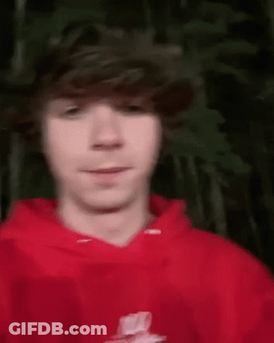 A Young Man Wearing A Red Hoodie Is Standing In Front Of A Tree . GIF