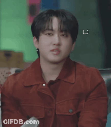 A Young Man Wearing A Red Jacket And Earrings Is Sitting On A Couch . GIF