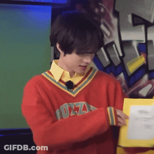 A Young Man Wearing A Red Sweater And A Yellow Shirt Is Holding A Piece Of Paper . GIF