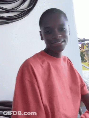 A Young Man Wearing A Red Sweatshirt Is Smiling While Sitting On A Balcony . GIF